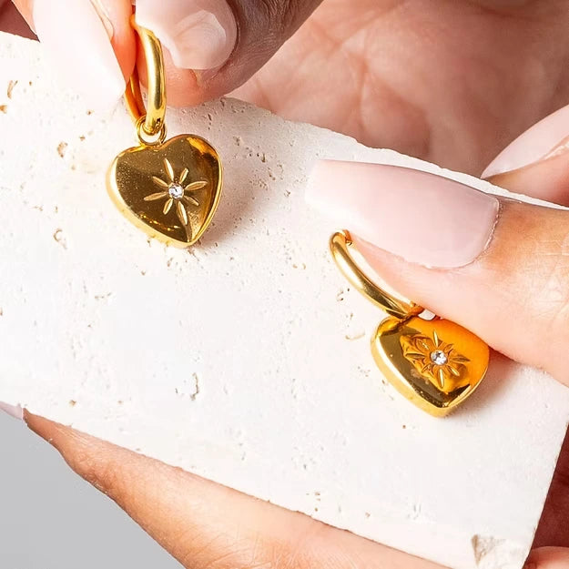 Gold heart-shaped earrings with floral designs held by a hand