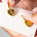 Gold heart-shaped earrings with floral designs held by a hand
